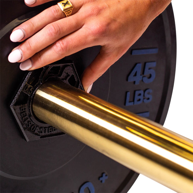 A hand, wearing a ring, adjusts a Fractional Iron Plate - 0.5 LB by Bells of Steel on an Olympic barbell; the hexagonal black grips of the 45 LB plate complement the barbell's metallic gold finish.