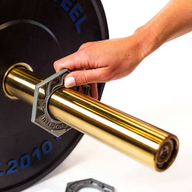 A hand securing a hexagonal weight collar on a gold Olympic barbell, featuring Bells of Steel's 0.5 LB Fractional Iron Plates with black cast iron design and blue text against a white background.