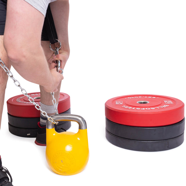 With focus on form, a person adjusts a chain linked to the yellow Bells of Steel Belt Squat Belt, readying for weighted dips. Nearby, stacks of red and black weight plates contrast against the white background. The individual sports black shorts and a gray shirt.