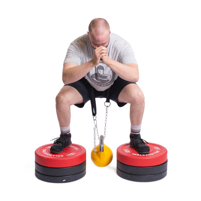 A person crouches with hands clasped, balancing on two stacks of red circular weights. A Bells of Steel yellow and black Belt Squat Belt is suspended by chains between the weights. They wear a gray shirt, black shorts, and black shoes.