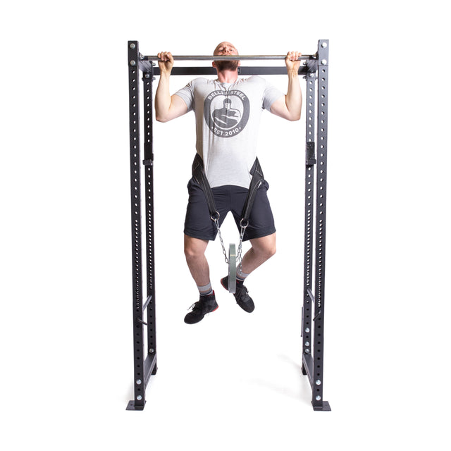 A man in a gray t-shirt and black shorts skillfully performs a pull-up on a sleek black metal rack using the versatile Belt Squat Belt by Bells of Steel, with a resistance band attached to his feet, showcasing strength and stability. This setup easily transitions to weighted dips or uses power rack attachments.