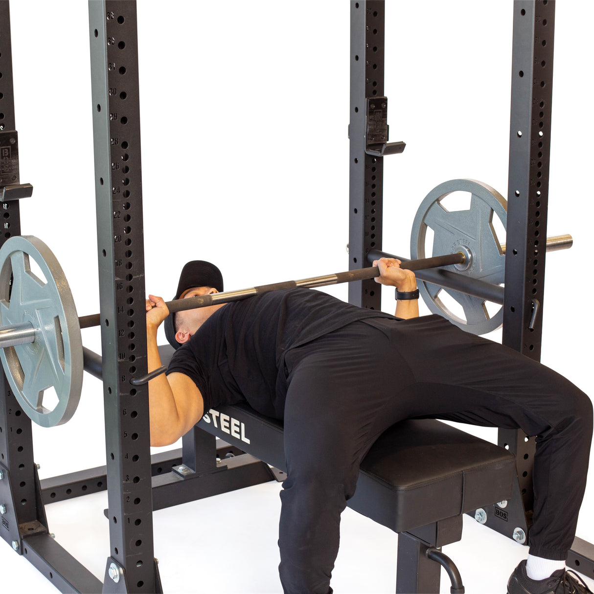 An individual in workout gear bench presses on a weight bench inside the Hydra Flat Foot Power Rack Builder by Bells of Steel. Modular pieces securely hold plates on both sides against a plain white backdrop.