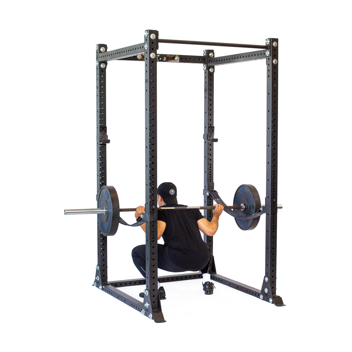 In a Hydra Flat Foot Power Rack Builder by Bells of Steel, a person in a black cap performs a rear-view squat using modular components and blue-weighted plates at both ends of the barbell. The rack is sleek and sturdy, with dimensions of 3" x 3" and ⅝" holes.