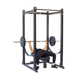 A person is performing a bench press inside the Bells of Steel Hydra Flat Foot Power Rack Builder (3" x 3", ⅝" Holes). They lie on a bench and hold a barbell with weights. The black power rack, equipped with adjustable safety bars, is set against a white background.