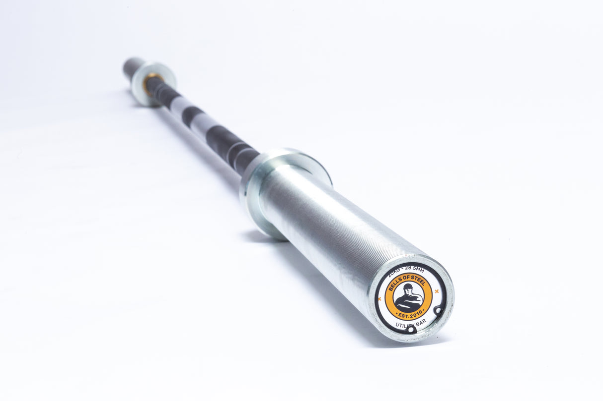 A close-up of the Bells of Steel Multi-Purpose Olympic Barbell shows its silver bar with black grip sections, set horizontally against a white background. The end cap has a bold yellow and black logo, ideal for powerlifting enthusiasts.