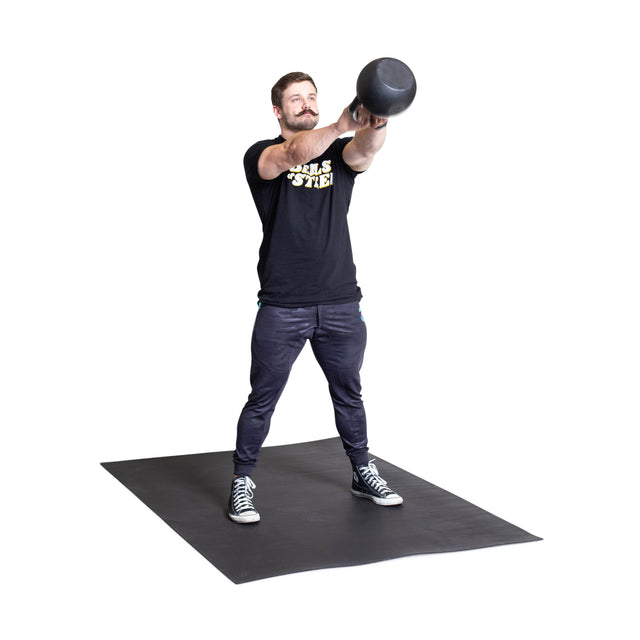 A man performs a kettlebell swing, lifting the weight to shoulder height on the Bells of Steel Premium Rubber Flooring 4' x 6', dressed in a black t-shirt, black pants, and gray sneakers.