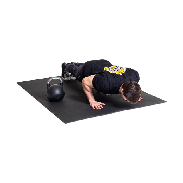 A man in black workout gear does push-ups on Bells of Steel Premium Rubber Flooring 4' x 6', with a kettlebell beside him. The white background highlights the setup and the durable recycled rubber surface.