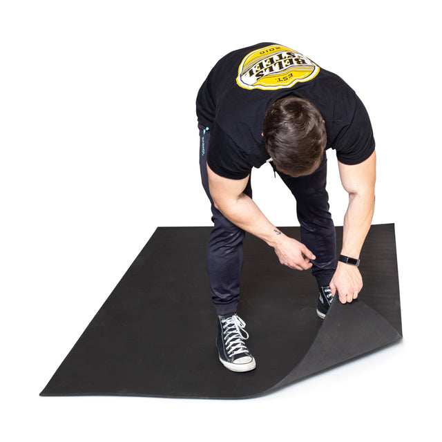 A person in a black t-shirt, dark pants, and sneakers bends down to lift the corner of a Bells of Steel Premium Rubber Flooring 4' x 6' mat.