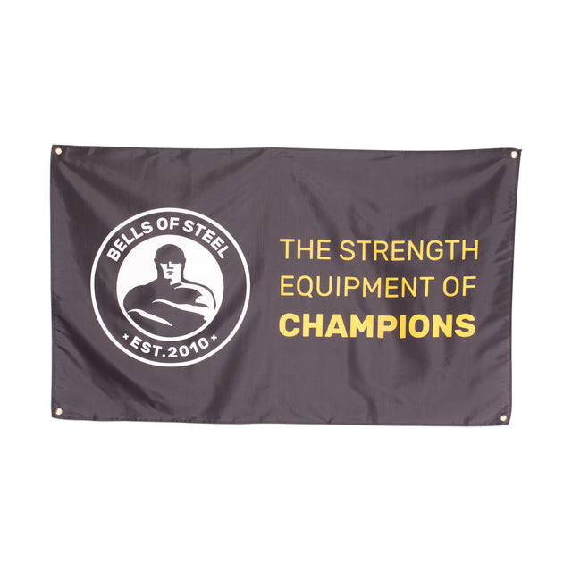 A black Bells of Steel flag features a circular logo with a flexing figure and "Bells of Steel Est. 2010" caption, alongside "The Strength Equipment of Champions" in bold gold letters, ideal for elevating your home gym decor.