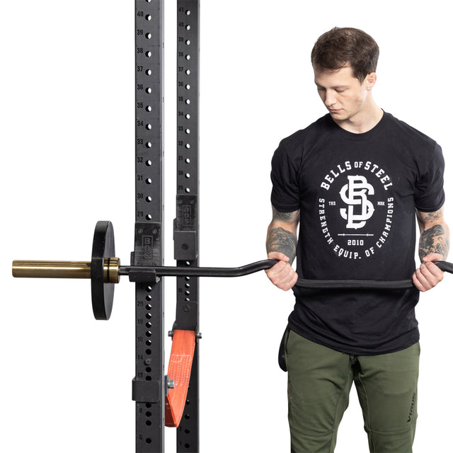 A person in a black "Bells of Steel" T-shirt and green pants is using a squat rack, concentrating on arm development with a partially loaded Rackable EZ Curl Bar.