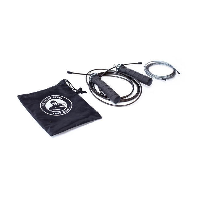 The Bells of Steel Jump Rope with ergonomic black and gray handles lies next to a black drawstring bag featuring the "Rope of Steel" circular logo, "Est. 2009." An adjustable cable is coiled nearby, ideal for boosting your cardio training routine.