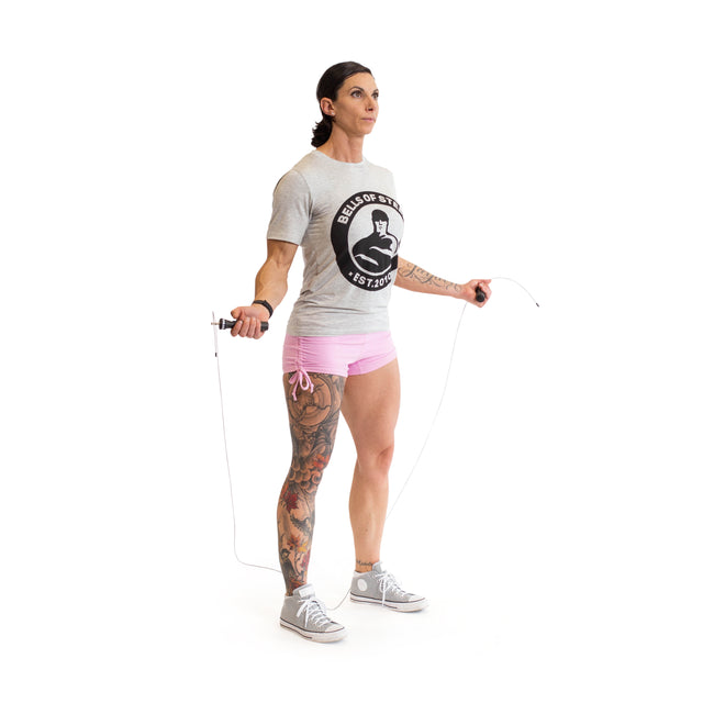 A person with short dark hair uses a Bells of Steel jump rope featuring ergonomic handles. They are dressed in a gray T-shirt, pink shorts, and gray high-top sneakers. One leg is adorned with colorful tattoos, and the plain white background highlights their focus on cardio training.