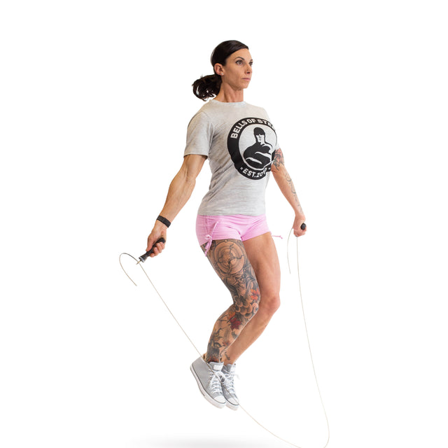 A tattooed person jumps rope with an adjustable Bells of Steel jump rope featuring ergonomic handles. They sport a gray logo t-shirt, pink shorts, and gray sneakers against a plain white background that highlights their cardio workout attire.