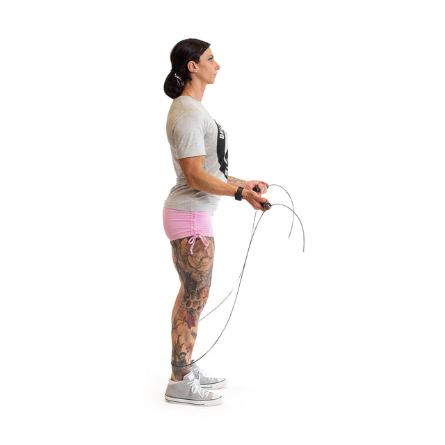 A person with long hair tied back wears a gray T-shirt, pink shorts, and gray sneakers while holding the Bells of Steel Jump Rope with ergonomic handles. They have sleeve tattoos on their left leg and arm against a plain white background, perfect for effective cardio training.