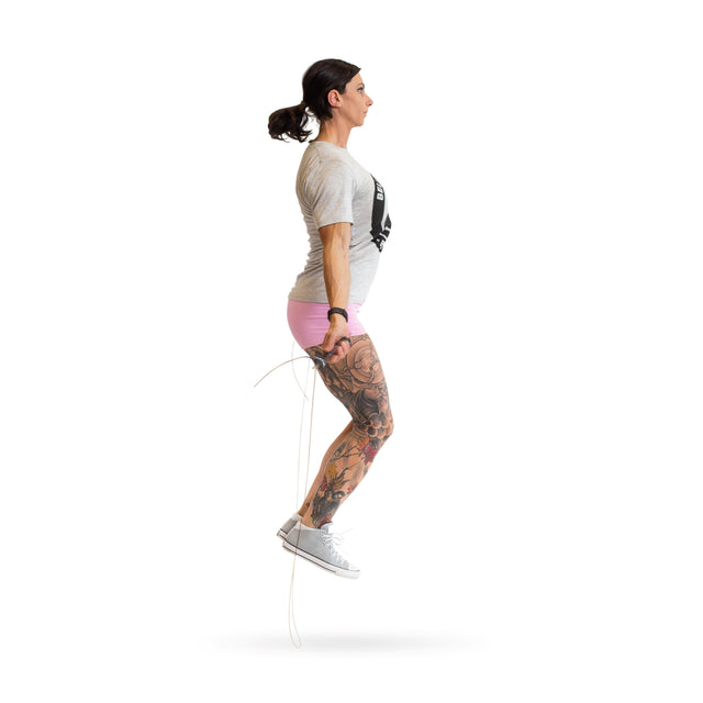 A person is mid-air, showcasing the Bells of Steel Jump Rope with ergonomic handles. They're in a gray T-shirt, pink shorts, and white sneakers. Their right arm gracefully holds the rope, while a tattoo on their left leg stands out against a plain white background—ideal for cardio enthusiasts.