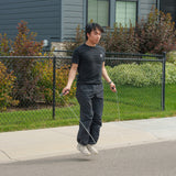 A person in a black shirt and pants uses the Bells of Steel Jump Rope for cardio training on a sidewalk in front of a house with a black chain-link fence and green bushes.