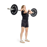 A woman powerlifts with the Juno Bar - Women's Utility Bar by Bells of Steel in a clean front rack position, demonstrating strength and technique. She's dressed in a black t-shirt, black shorts, and white sneakers against a simple white background.