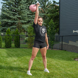 A person stands on grass, holding a pink Bells of Steel Competition Kettlebell overhead with one arm, dressed in a black t-shirt, black shorts, and white sneakers. Trees and a fence appear in the background—demonstrating how identical-sized Bells of Steel Competition Kettlebells help maintain training consistency across various weights.