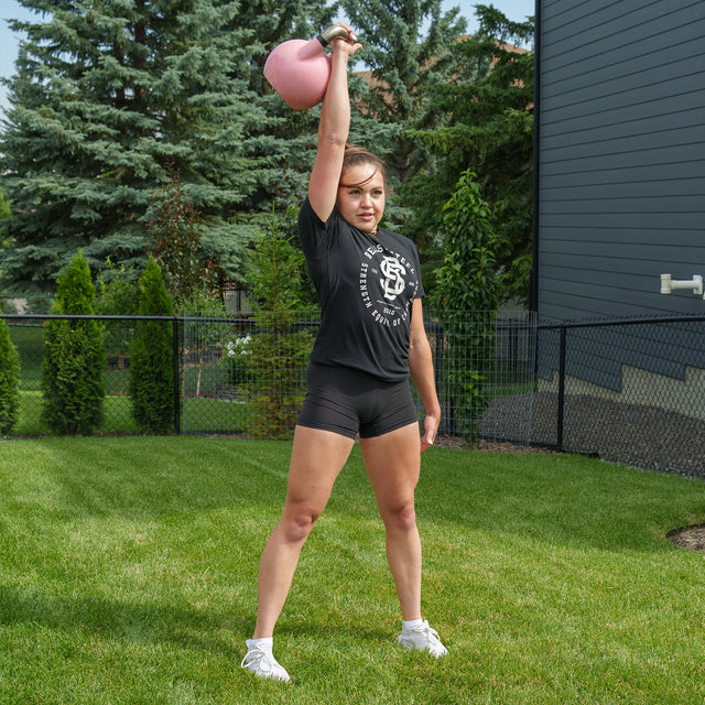 A person stands on grass, holding a pink Bells of Steel Competition Kettlebell overhead with one arm, dressed in a black t-shirt, black shorts, and white sneakers. Trees and a fence appear in the background—demonstrating how identical-sized Bells of Steel Competition Kettlebells help maintain training consistency across various weights.