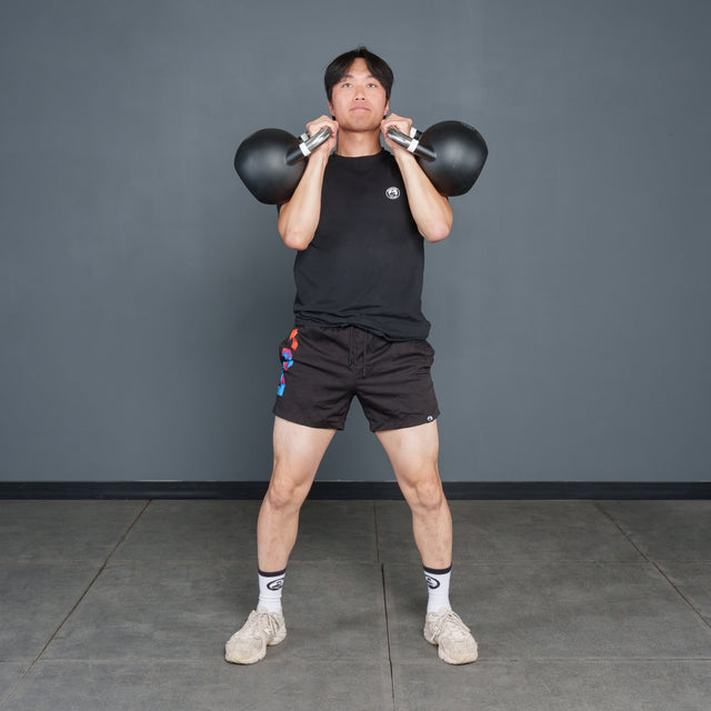 A person wearing a black shirt, black shorts, white socks, and sneakers stands on a gym floor, holding two Bells of Steel Competition Kettlebells at shoulder height, preparing to exercise.