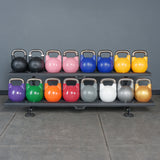 Two shelves display Bells of Steel Competition Kettlebells, each with a metal handle and a unique color for every size, all neatly arranged against a gray wall on a tiled gym floor.