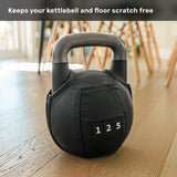 A Bells of Steel USA Kettlebell Wrap in stitched leather, labeled “125,” protects a black competition kettlebell sitting on a wooden floor in a sunlit room. Text above reads: “Keeps your kettlebell and floor scratch free.”.