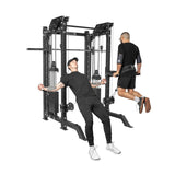 Two men exercise with THE KOLOSSOS - Smith Functional Trainer by Bells of Steel; one is doing tricep pushdowns, the other assisted chin-ups using a weighted belt. They're both wearing black athletic clothing suited for their home gym setup.