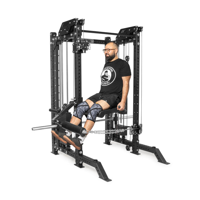 Wearing a black shirt, shorts, and knee pads, a person works out on THE KOLOSSOS - Smith Functional Trainer by Bells of Steel. They perform a seated leg exercise using the machine's sleek black pulleys and bars to enhance their strength training.