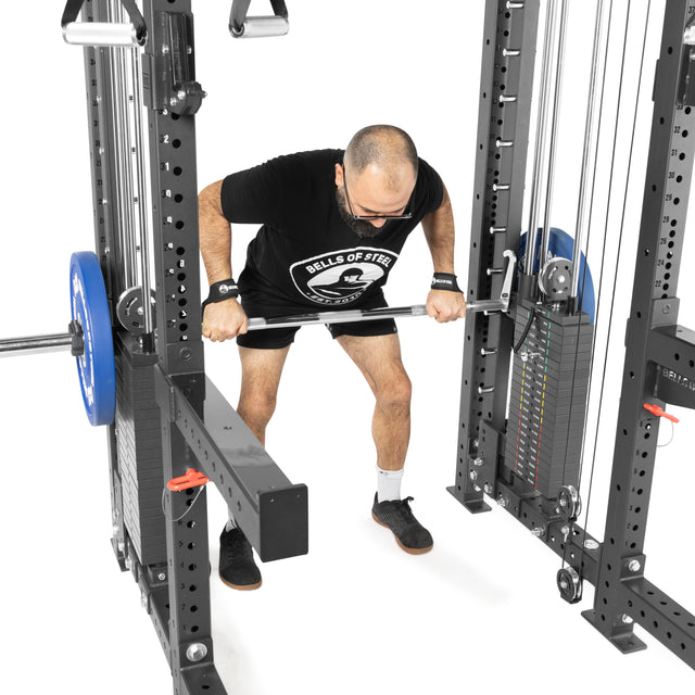 Wearing a black shirt and shorts, a person uses THE KOLOSSOS - Smith Functional Trainer from Bells of Steel to perform a bent-over row in the gym, focusing on form while gripping the bar near the power rack and weight stacks. The machine includes blue weight plates.