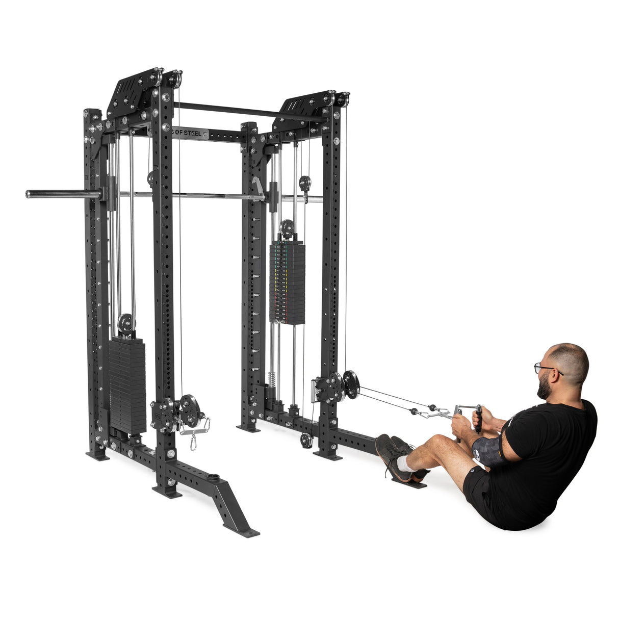 A man in a black shirt and shorts uses THE KOLOSSOS - Smith Functional Trainer by Bells of Steel for a seated row exercise, pulling handles with focused intensity.