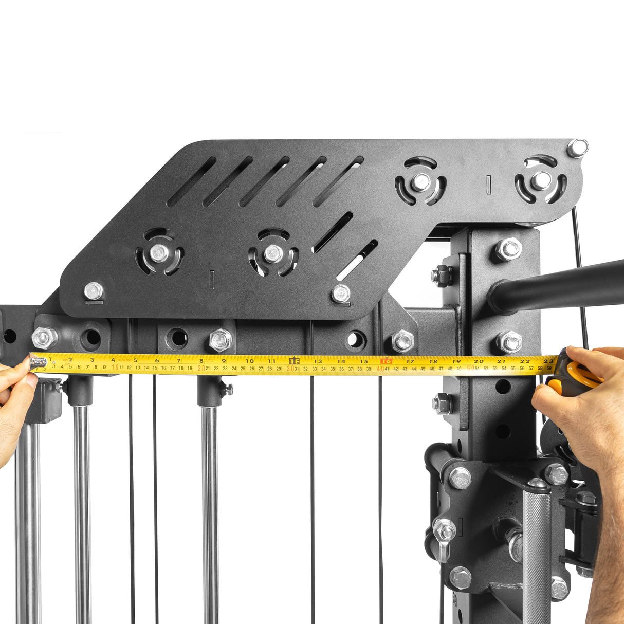 A person uses a yellow tape measure to check the width of THE KOLOSSOS - Smith Functional Trainer, a black metal frame with pulleys and cables, from Bells of Steel, holding the tape measure at both ends.