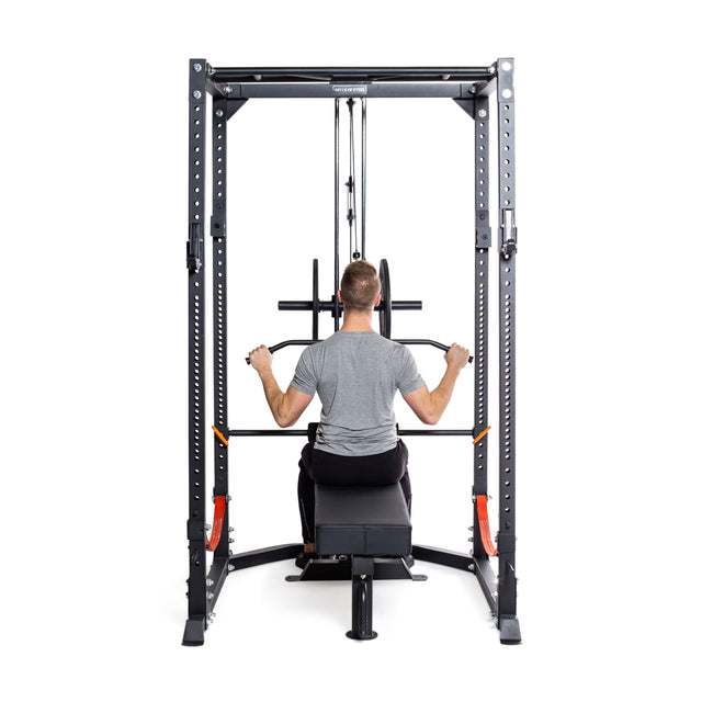 A person in a gray T-shirt and black pants uses the Bells of Steel Rack Lat Pulldown/Row Attachment. They pull down on the bar with both hands on this light commercial/residential power rack, set against a white background.