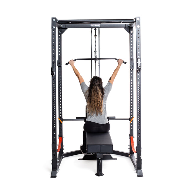 A person with long hair sits on a workout bench using a Bells of Steel Rack Lat Pulldown / Row Attachment. The metallic frame features an adjustable pulley system and multiple weight options, suitable for light commercial or residential power racks.