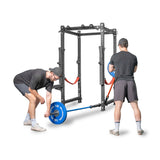 Two individuals in athletic wear and caps are preparing for a strength workout with a blue barbell in the squat rack, which is equipped with orange safety straps and the "Bells of Steel" Landmine Rack Attachment, surrounded by various exercise equipment.