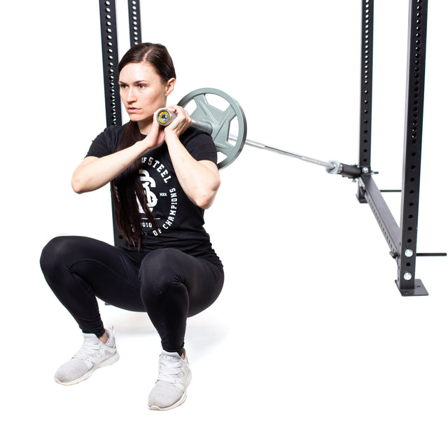 In a gym, someone with long brown hair, dressed in a black T-shirt, black leggings, and white sneakers, is using the Bells of Steel Landmine Rack Attachment for a front squat. The squat rack enhances this dynamic workout against the pristine white floor backdrop.