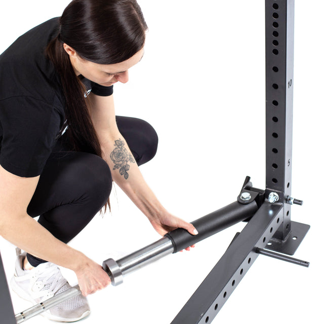 A person with long dark hair, wearing a black t-shirt, black leggings, and white sneakers, kneels on the ground attaching a metal bar to a Bells of Steel Landmine Rack Attachment. Their forearm features a floral tattoo.