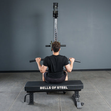 In a gym, a person on a Bells of Steel bench faces a cable machine and grasps the Bells of Steel Fat Bar - Lat Pulldown Bar, ready to perform lat pulldowns to increase grip strength.