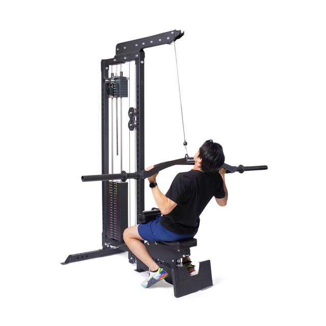 A person in a black shirt and blue shorts uses the Bells of Steel Lat Pulldown Low Row Machine, pulling its black bar down to their chest with a weight stack in a gym setting.