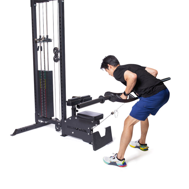 Wearing a black shirt and blue shorts, a person performs a seated row with focused precision, drawing the handle of the Bells of Steel Lat Pulldown Low Row Machine towards their chest amidst its weights and pulleys in a gym setting.