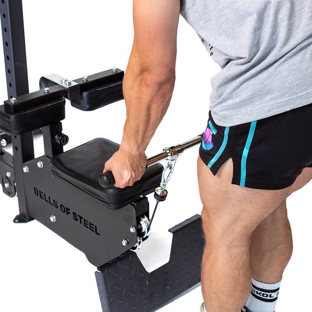 A person in a gray shirt and black shorts uses a Bells of Steel Lat Pulldown Low Row Machine, gripping the handle and positioning themselves on the black padded seat, ready for exercise.