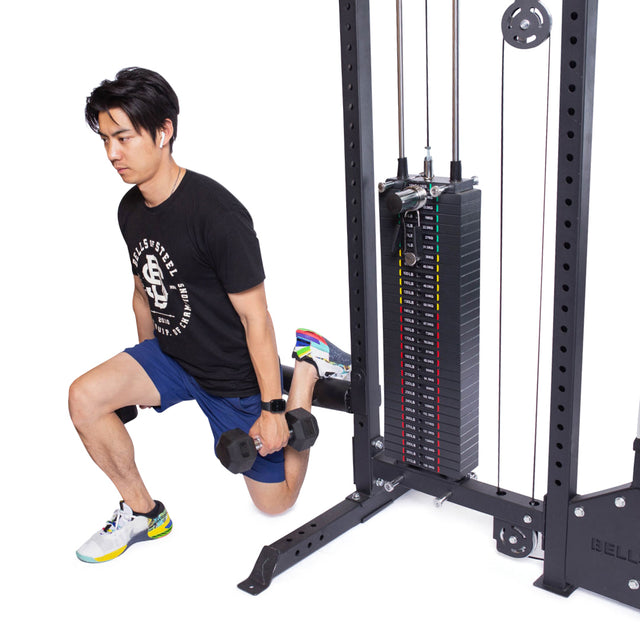 Using a Bells of Steel Lat Pulldown Low Row Machine in their home gym, a person performs a split squat with an elevated foot on the frame. Holding dumbbells in each hand, they are dressed in a black t-shirt, blue shorts, and sneakers.