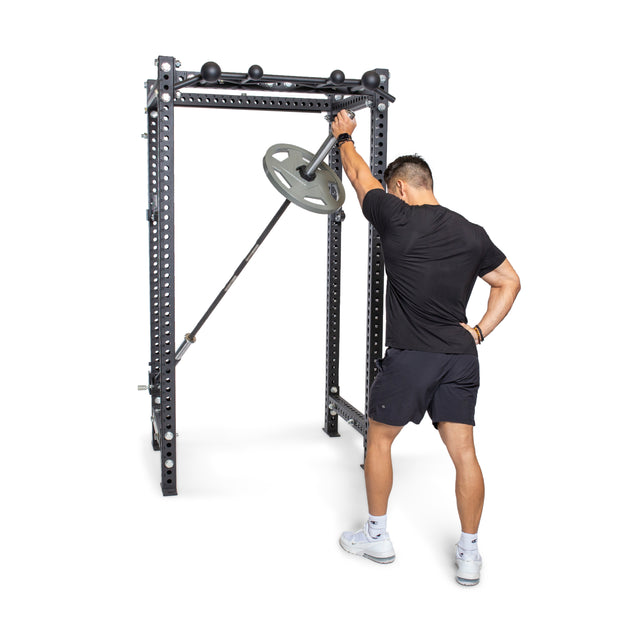 A person in a black shirt and shorts performs a landmine exercise with the Bells of Steel Landmine Rack Attachment, gripping the barbell with both hands and pushing the weighted end while facing the power rack.