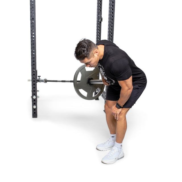 In a gym, using the Bells of Steel Landmine Rack Attachment, a person bent slightly forward with feet shoulder-width apart performs a bent-over barbell row. They're dressed in black with white shoes against the backdrop of a gym rack, enhancing their dynamic workout.