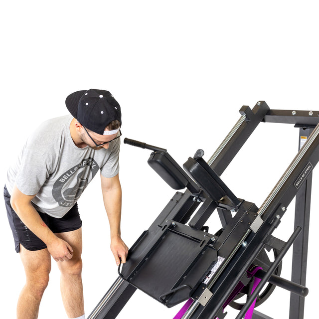 Wearing a gray t-shirt, black shorts, cap, and glasses, a person adjusts the Bells of Steel Leg Press Hack Squat Machine. This sleek black-framed equipment with multiple handles features a slanted seat ideal for hack squats against a white background.