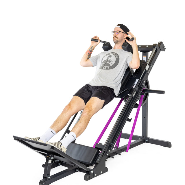 In their home gym, a person wearing glasses and a cap uses the Bells of Steel Leg Press Hack Squat Machine. Dressed in a gray shirt, black shorts, and sneakers, they fit into the minimalist setup with its black seat, purple accents, and plain white background.