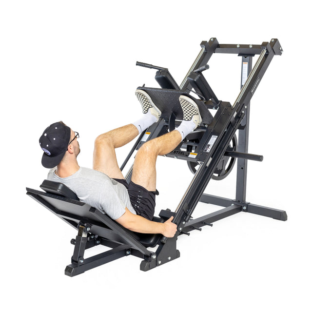 A person outfitted in a black cap, glasses, gray shirt, and black shorts is using the Bells of Steel Leg Press Hack Squat Machine in their home gym. They are lying back on the machine's seat, pushing against its footplate with their feet.