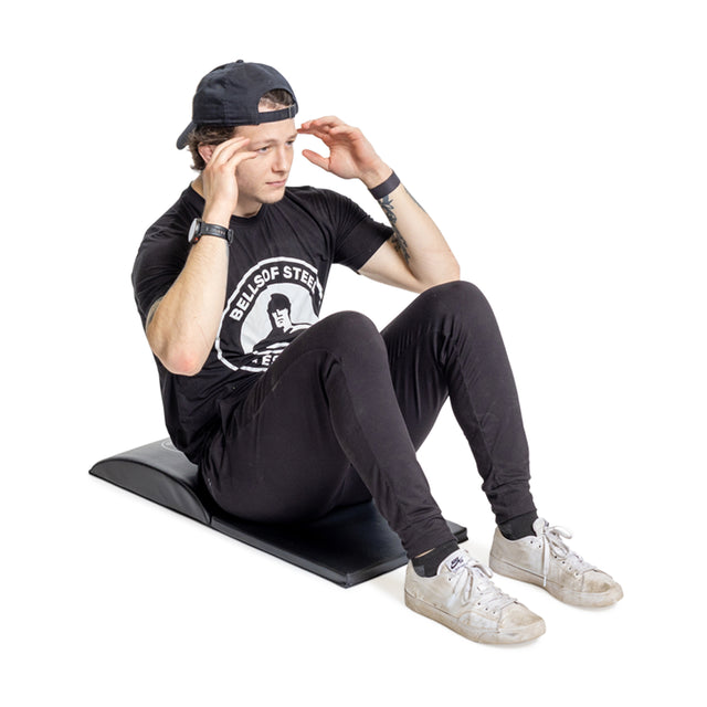 A person in a black cap, shirt, and pants does a sit-up on a Bells of Steel Luxury Sit Up Mat. With hands at their temples and white sneakers on, they concentrate intently on their core workout against a plain white background.