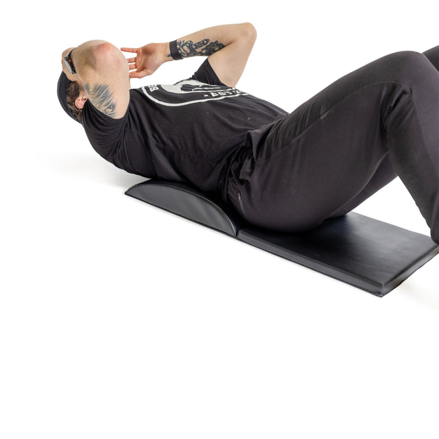 A person lies on a Bells of Steel Luxury Sit Up Mat, performing core workouts in black T-shirt and pants. With hands behind their head and elbows bent, a tattoo shows on one arm as they maintain proper lumbar curvature.