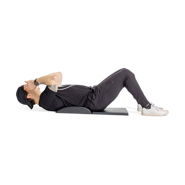 In a bright space, a person in athletic wear—cap, T-shirt, pants, sneakers—performs core workouts on a Bells of Steel Luxury Sit Up Mat. Maintaining perfect form with bent knees and hands behind their head, they ensure lumbar curvature during the exercise.