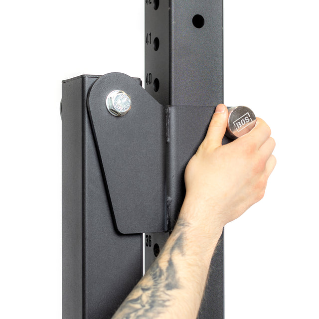 A person with a tattooed arm uses a wrench to tighten a bolt on a black metal power rack, which features numbered holes for adjustable height and utilizes Bells of Steel Magnetic Rack Attachment Pins for efficient security.